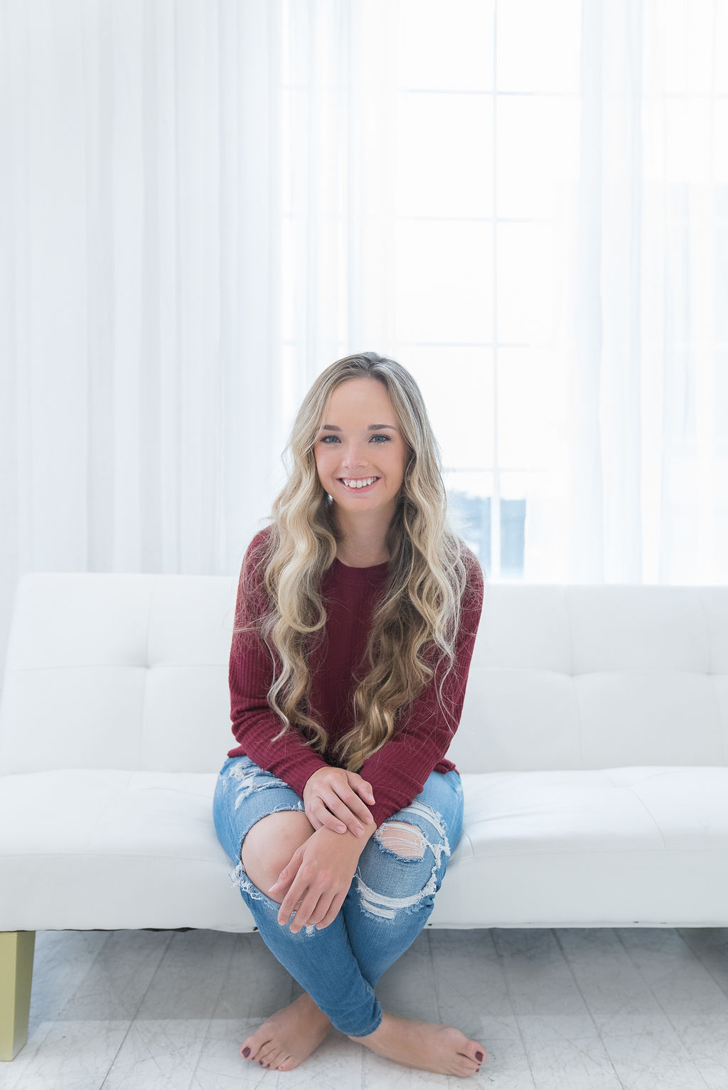 image of Amber Broder sitting on couch and smiling at camera wearing red sweater and ripped jeans