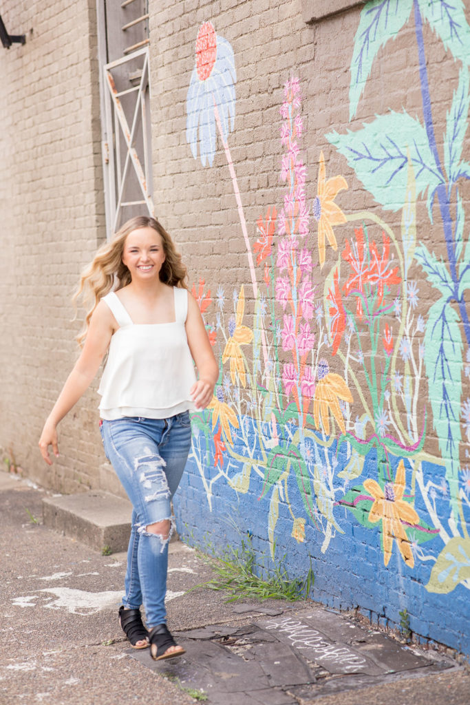 girl walking down the street in white top and jeans next to mural on brick wall